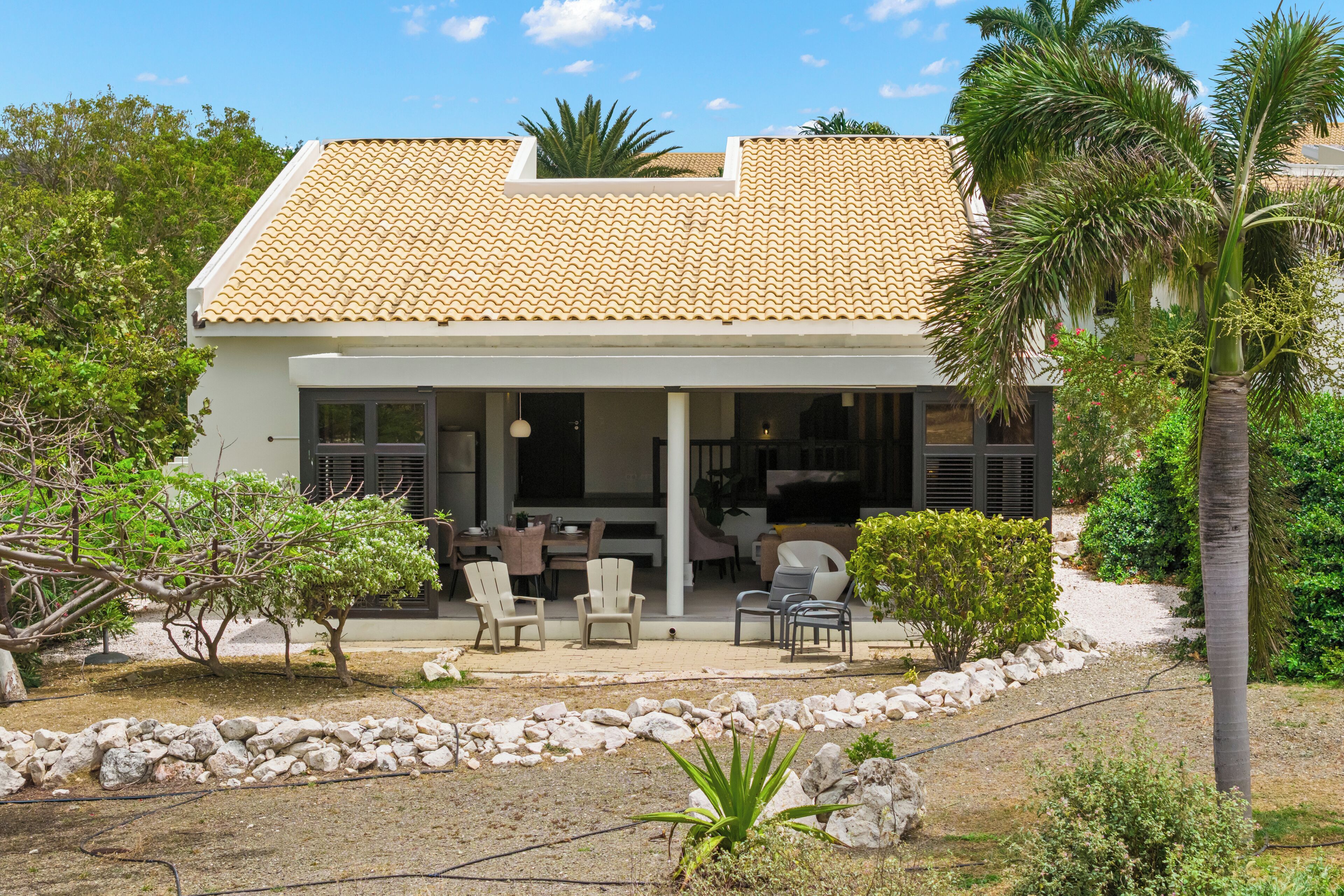 Ein zweistöckiges Haus mit Ziegeldach, einer überdachten Terrasse mit Sitzgelegenheit und einem Garten mit Steinen und Pflanzen.