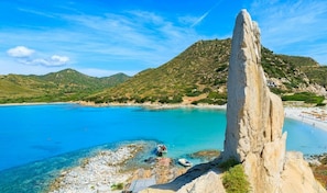 A coastal landscape with a prominent rock formation, clear blue waters, and a hilly backdrop.