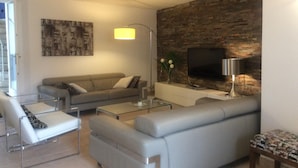 A modern living room with a grey sofa, a glass coffee table, and a TV mounted on a brick wall.