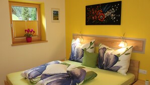 Ein Schlafzimmer mit einem Bett, einem Nachttisch, einem Fenster mit Jalousien, einer Zimmerpflanze und einem Bild an der Wand.