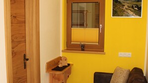 Ein Zimmer mit Holztür, braunem Ledersofa, einem kleinen Holzregal und einem Fenster mit Jalousien.