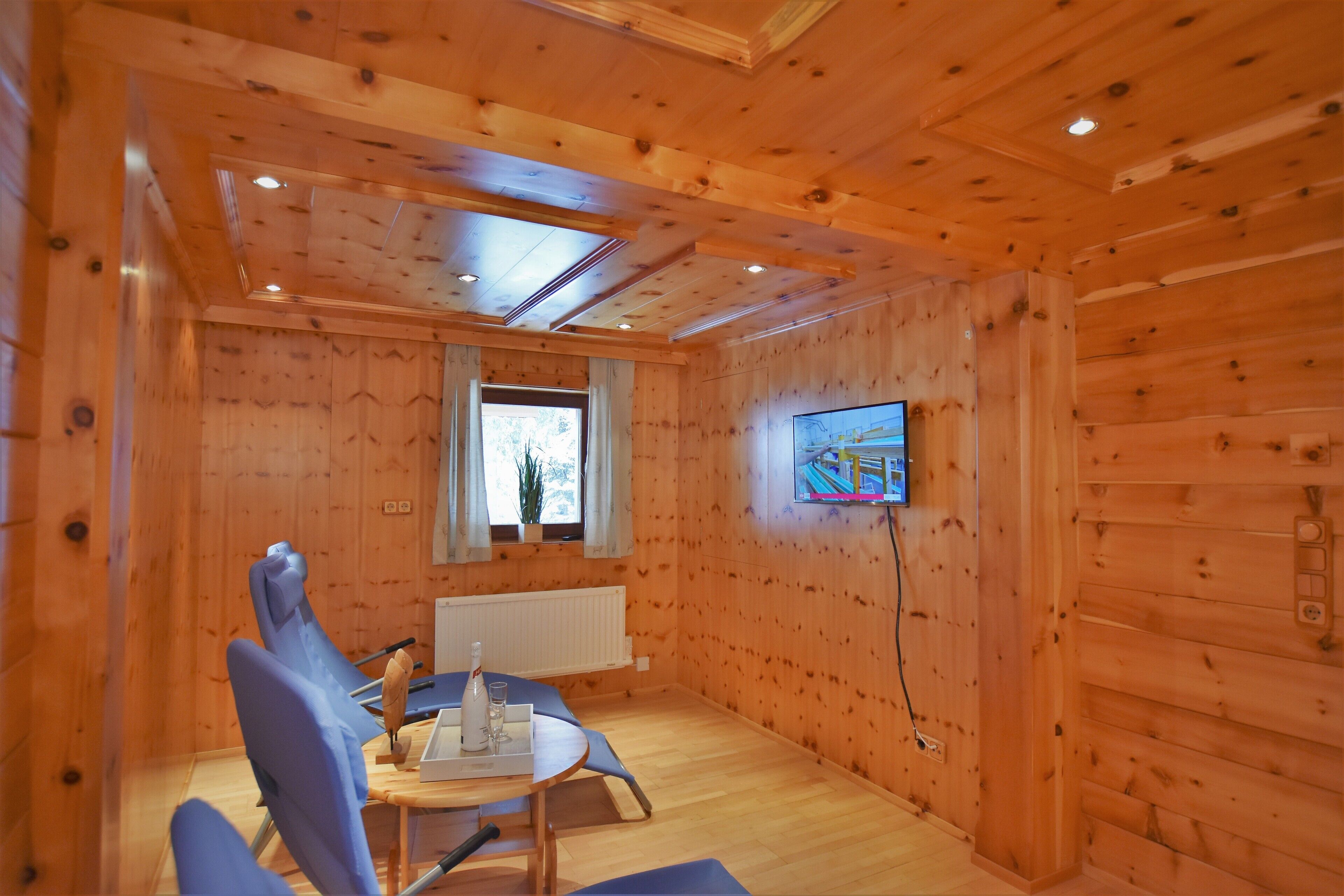 Una habitación con paneles de madera, un televisor fijado a la pared, una mesa con sillas y un ventanal que proporciona luz natural.