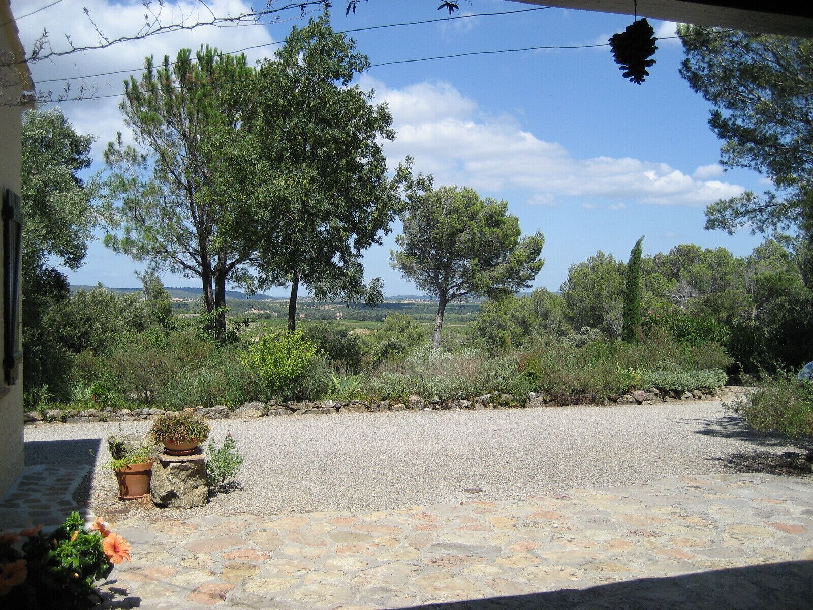 Un camino de grava conduce a una casa con un patio de piedra, rodeada de árboles y un cielo despejado.