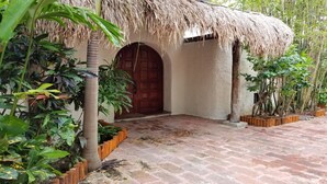 Un edificio tradicional con techo de paja y puerta de madera, rodeado de abundante vegetación y plantas en macetas.