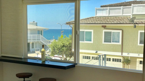 A room with a large window overlooking a coastal area, featuring a balcony with a view of the sea and adjacent buildings. - Steps to sand! Ocean view, Walk to village, family friendly, A/C! (Carlsbad)