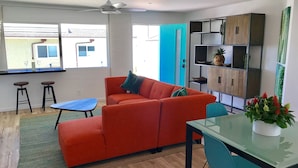 A modern living room with a red sofa, a glass coffee table, and a blue accent table. There are two bar stools and a small table with a potted plant. A teal cabinet with shelves is against the wall, and a door with a teal frame is visible. - Steps to sand! Ocean view, Walk to village, family friendly, A/C! (Carlsbad)