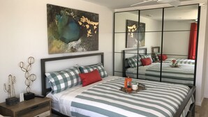 A modern bedroom with a large bed, striped bedding, a nightstand, a mirror, and abstract wall art. - Steps to sand! Ocean view, Walk to village, family friendly, A/C! (Carlsbad)