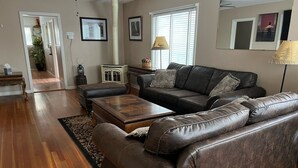 Ein Wohnzimmer mit braunem Ledersofa, einem Holztisch und einem gemusterten Teppich. Eine Lampe steht auf einem Beistelltisch, und ein Fenster mit Jalousien ist vorhanden.