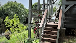 A wooden staircase leading up to a covered porch with a metal railing, surrounded by lush greenery and flowering plants. - King en suites.  Enjoy panoramic views of city and mountains, Walkable to town. (Asheville)
