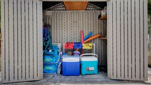 A small shed with a corrugated metal exterior and a wooden roof. Inside, there are two coolers, a stack of folded clothes, and various items including a blue bag and a red and yellow tube. - Tana’s Siesta Getaway - Heated Private Pool (Sarasota)