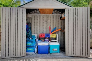 A small shed with a corrugated metal exterior and a wooden roof. Inside, there are two coolers, a stack of folded clothes, and various items including a blue bag and a red and yellow tube. - Tana’s Siesta Getaway - Heated Private Pool (Sarasota)
