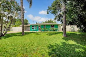 A single-story house with a green exterior, surrounded by a well-maintained lawn and palm trees.