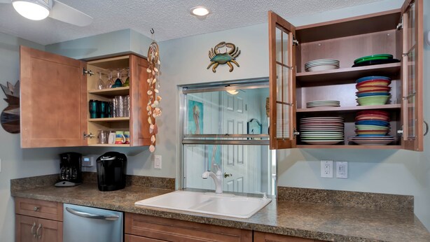 A kitchen with a granite countertop, stainless steel dishwasher, wooden cabinets, a coffee maker, and a glass-fronted cabinet with plates. - Tana’s Siesta Getaway - Heated Private Pool (Sarasota)