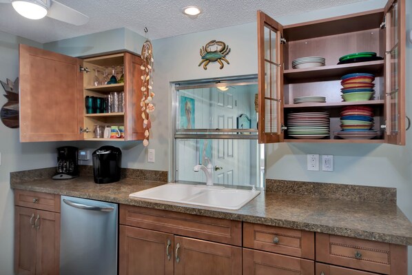 A kitchen with a granite countertop, stainless steel dishwasher, wooden cabinets, a coffee maker, and a glass-fronted cabinet with plates.
