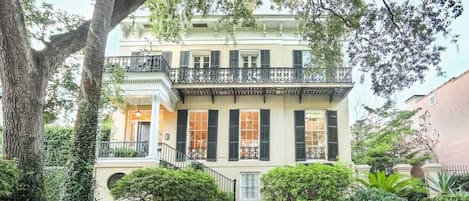 Stately building with a central entrance, flanked by windows and a balcony with wrought iron railing, set amidst lush greenery.