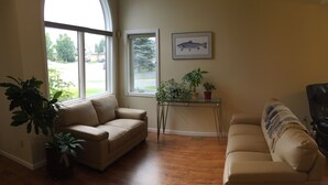 Un salon avec deux canapés beige, une table basse en verre ornée de plantes, un tableau représentant un poisson encadré accroché au mur, et une vue sur une rue de banlieue à travers de grandes fenêtres.