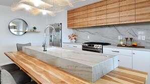 A modern kitchen with a marble countertop, wooden cabinets, stainless steel appliances, and a unique pendant light fixture.