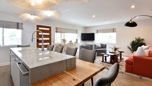 A modern kitchen with a marble island, grey chairs, and a wooden dining table. There is a sofa and a TV in the living area.
