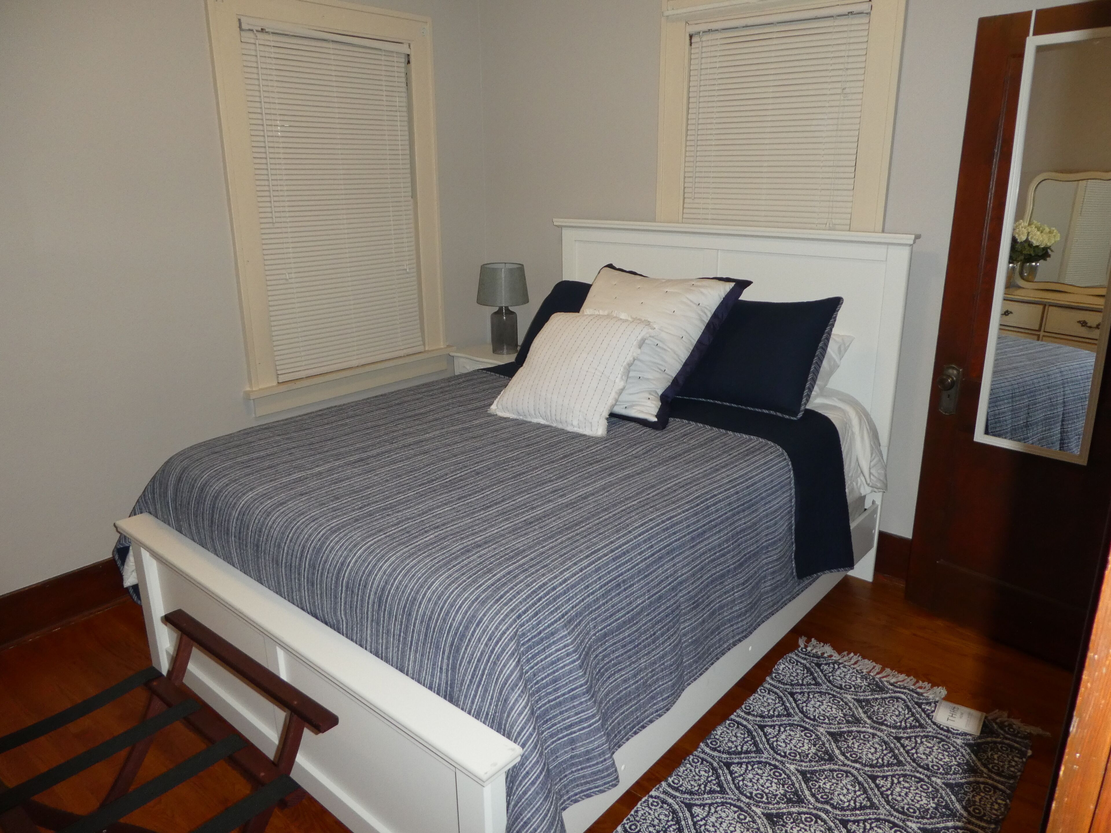 Un dormitorio con cama, dos ventanas con persianas, una mesita de noche con lámpara, una silla de madera, alfombra estampada y un espejo en la pared.