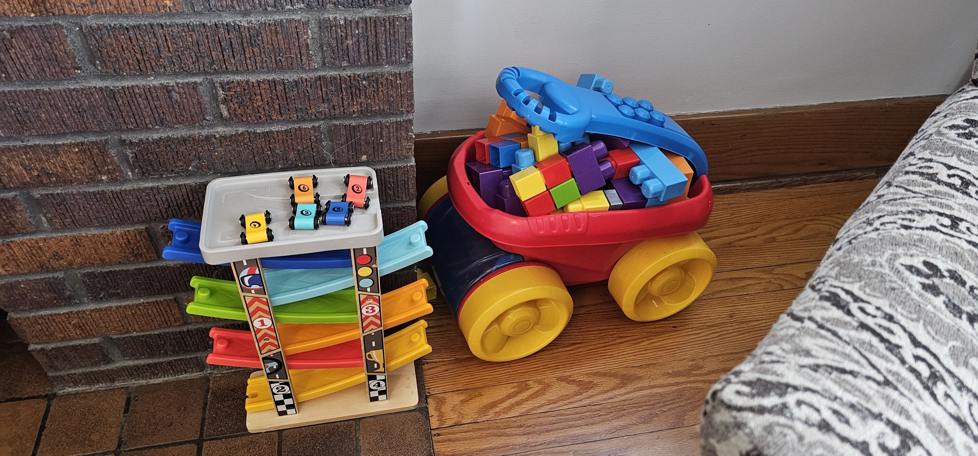 A toy train set with colorful blocks and a toy car on a wooden floor next to a brick wall.