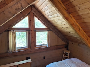 Une cabane en bois avec un plafond en pente, deux fenêtres avec des rideaux, un petit tabouret et un lit.