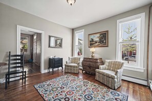 A living room with hardwood flooring, a patterned rug, a sofa, a chair, a small table, and a lamp. There are two windows with blinds, a door leading to another room, and a ceiling light fixture. - Culp House: Ideal Location, Historic Gettysburg! Can Rent with the Cricket House (Gettysburg)