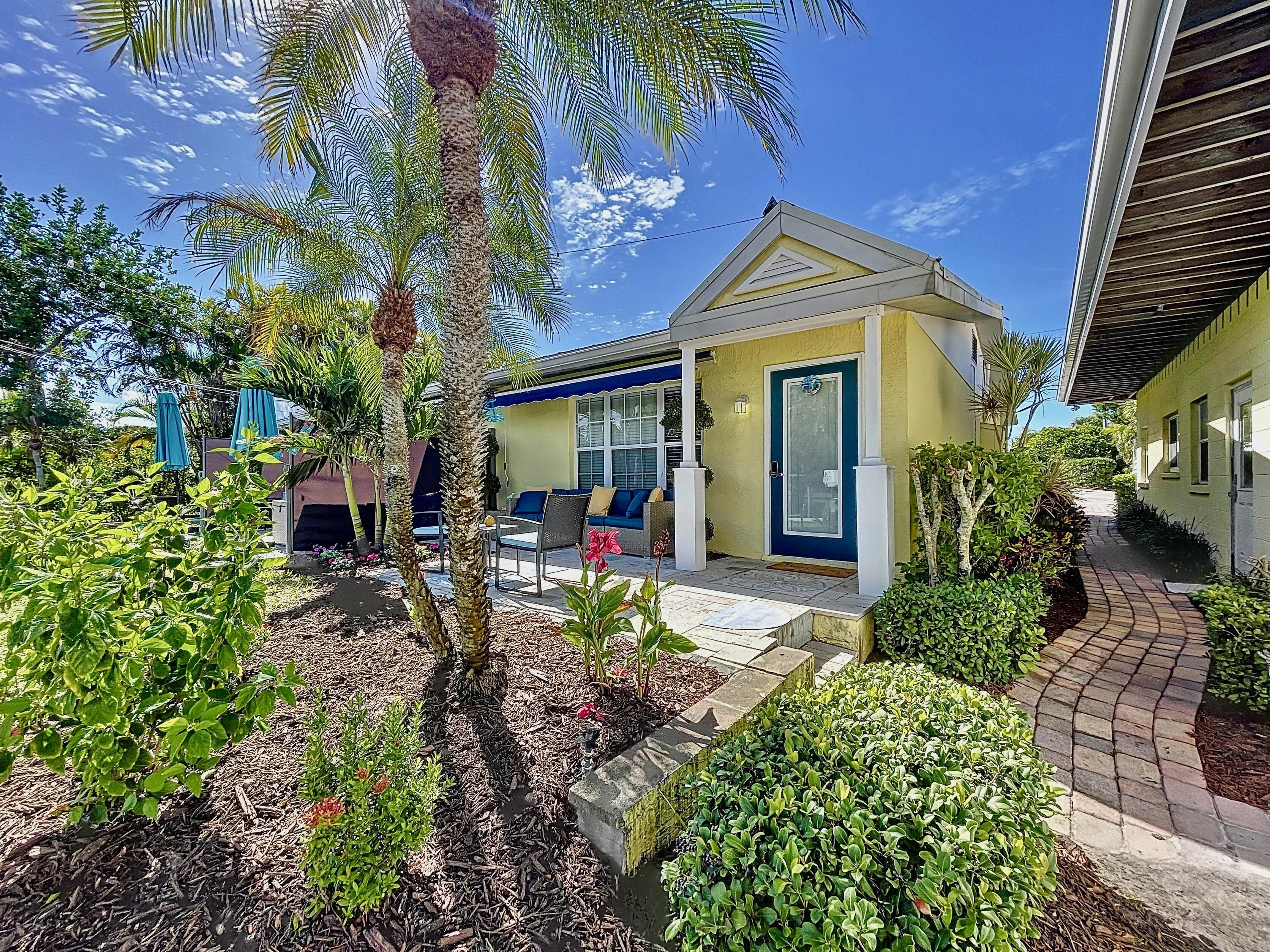 A residential house with a blue door, a covered patio, and a small garden area with palm trees and shrubs.