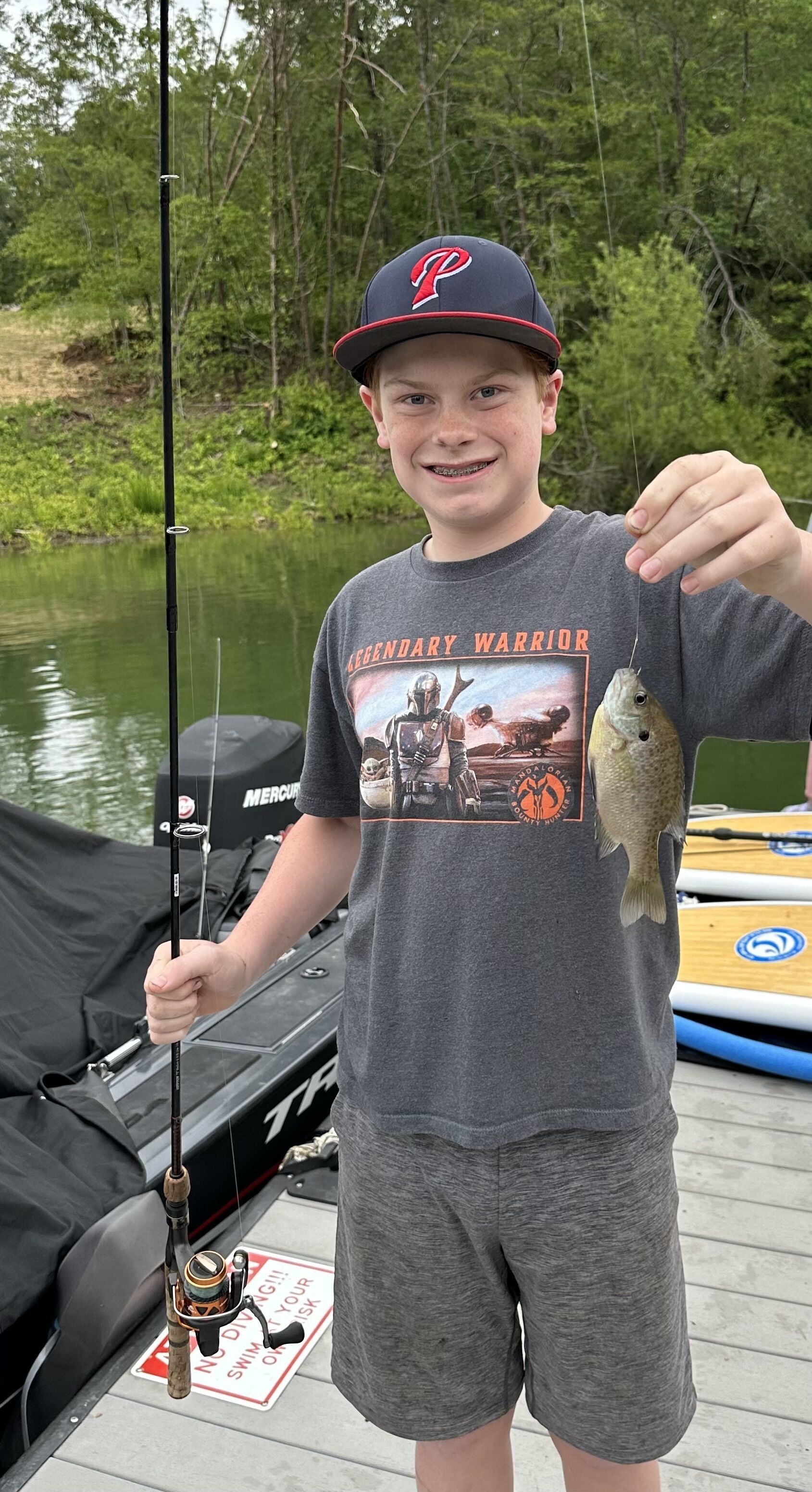 A person holding a fish and a fishing rod on a dock with boats in the background.