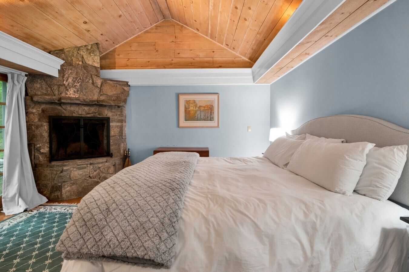 A bedroom with a stone fireplace, a bed with white bedding, and a wooden ceiling.