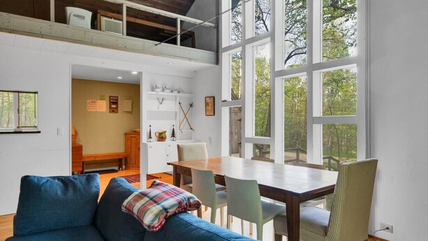 Een gezellige woonkamer met een blauwe bank, een houten eettafel en stoelen. Er is een groot raam met uitzicht op bomen.