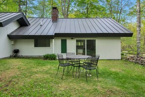 A single-story house with a metal roof, a brick chimney, a sliding glass door, and a patio with a table and chairs. - A-Frame Chalet | Floor-to-Ceiling Windows, 1 Mi to Bromley (Peru)