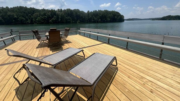 Una terraza de madera con sillones de descanso y una mesa, con vista a un lago tranquilo y una isla boscosa en la distancia.
