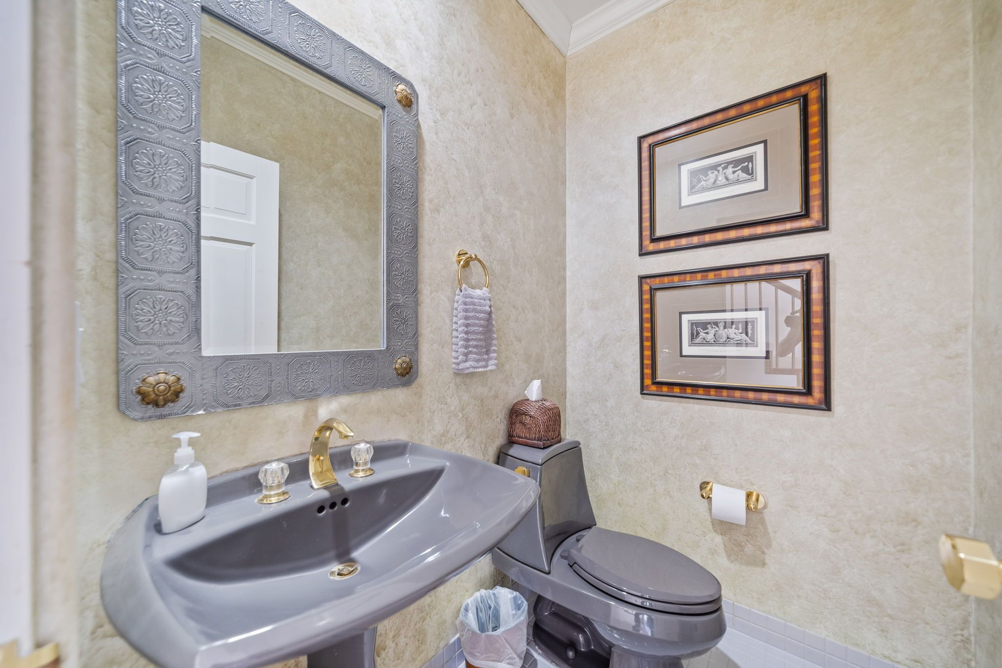 A bathroom with a grey sink, a toilet, a large mirror with ornate detailing, and two framed pictures on the wall.