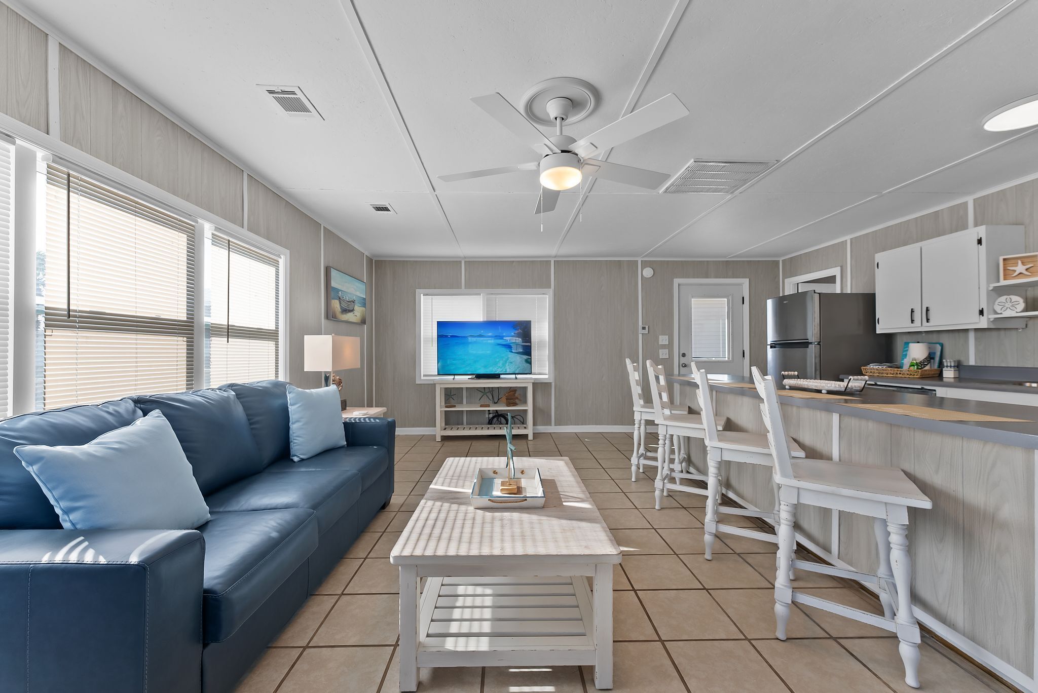 A modern living room with a blue sofa, a coffee table, and a TV. There is a kitchen area with white cabinets and a refrigerator.