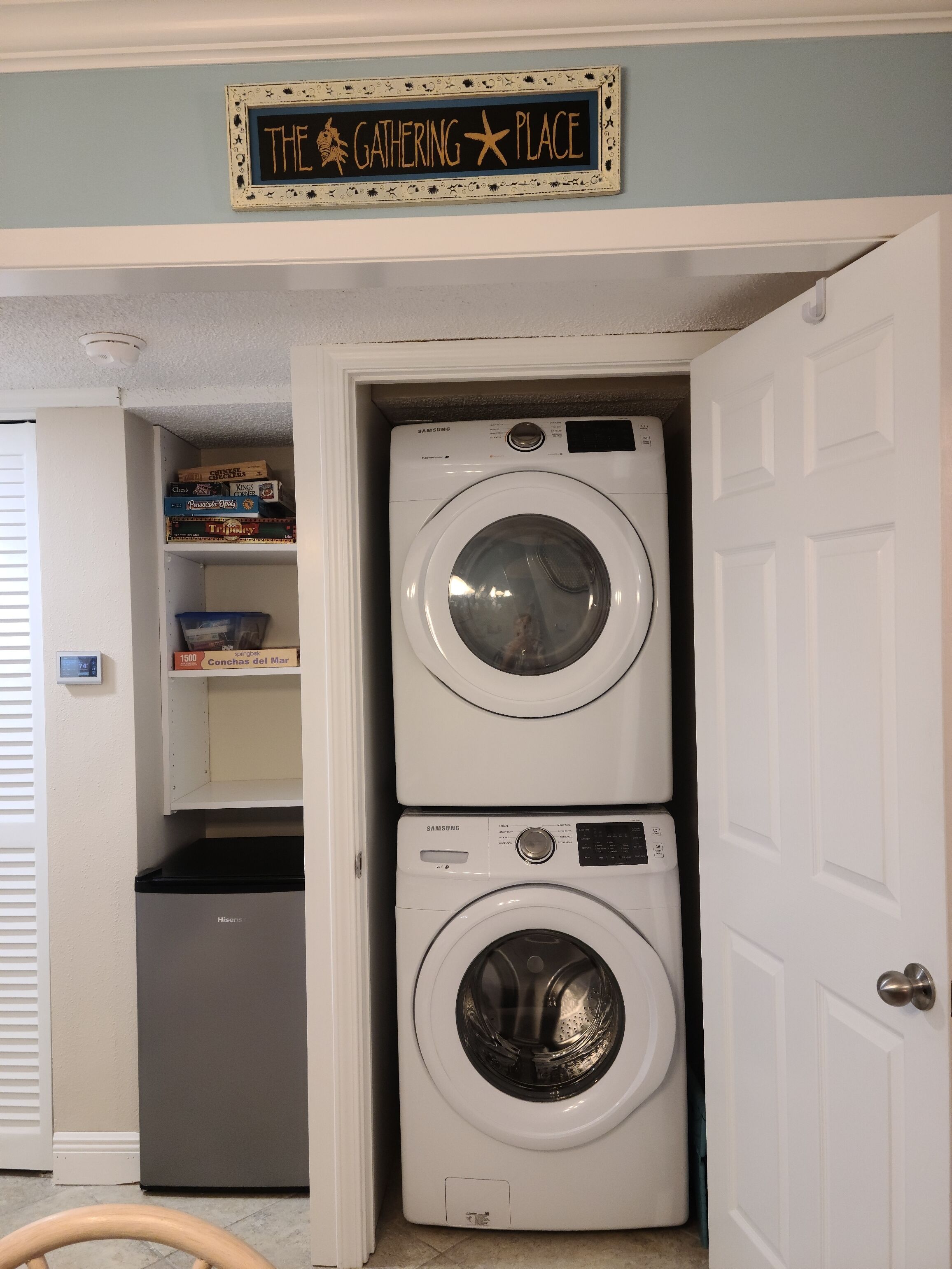 A laundry room with two stacked front-loading washers and dryers, a shelf with board games, and a sign above reading "THE GATHERING PLACE".