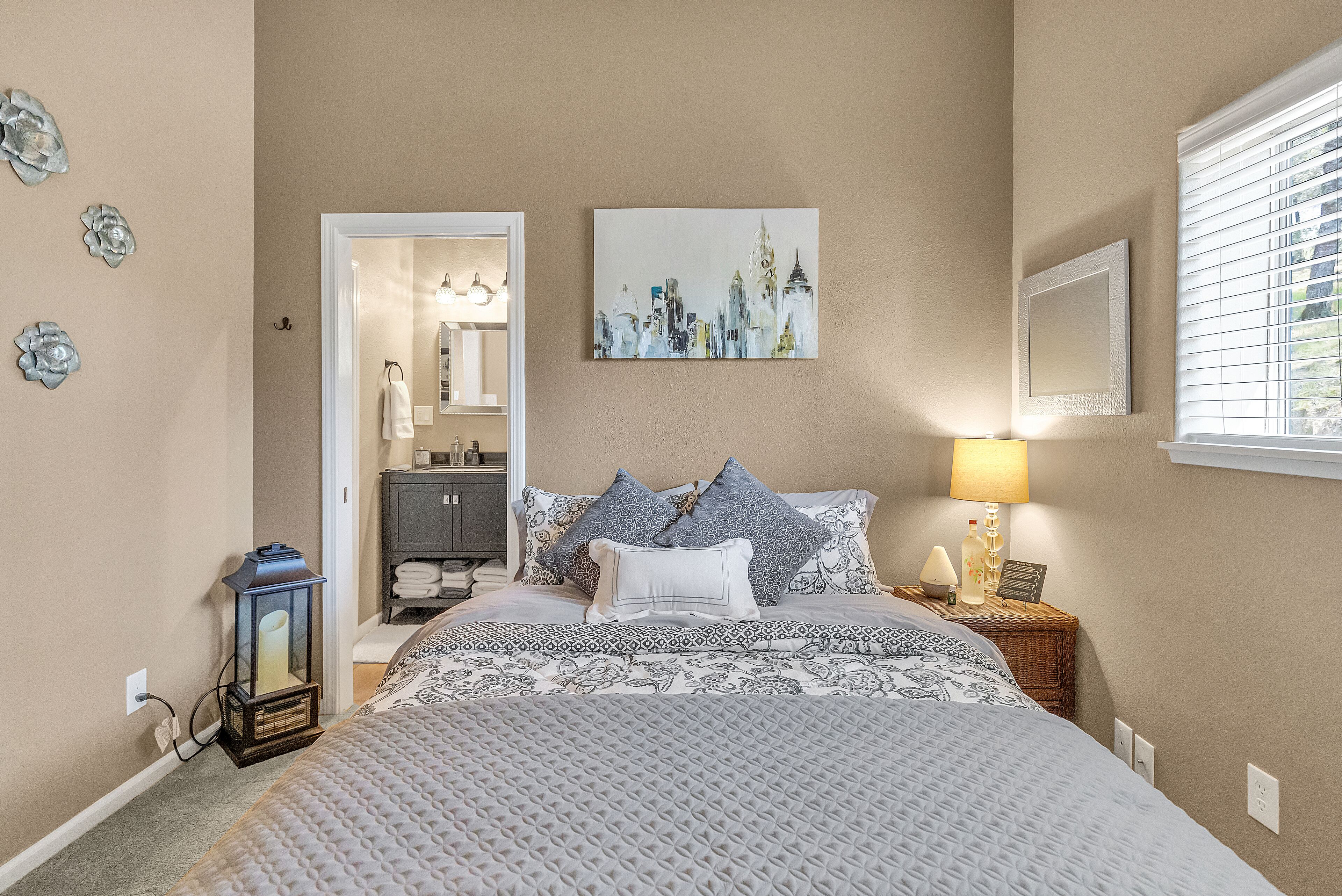 Une chambre à coucher avec un lit, une table de chevet, une lampe, un miroir et un tableau représentant un paysage urbain.