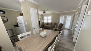 Un comedor con mesa de madera y sillas blancas, una cocina con refrigerador, y una sala de estar con sofá y un perro.