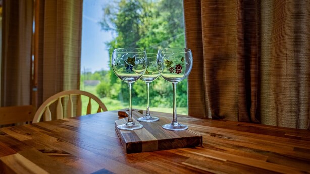 A wooden table with three wine glasses on a wooden board, positioned near a window with a view of greenery.
