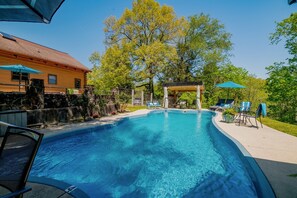 Ein klares, blaues Schwimmbecken mit Liegestühlen und Sonnenschirmen, umgeben von Bäumen und einer Steinmauer.