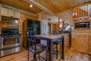 Ein Holzblockhaus-Interieur mit Küchenzeile, inklusive Herd, Backofen und Mikrowelle, einem Esstisch mit Stühlen und einer Treppe zu einer oberen Ebene.