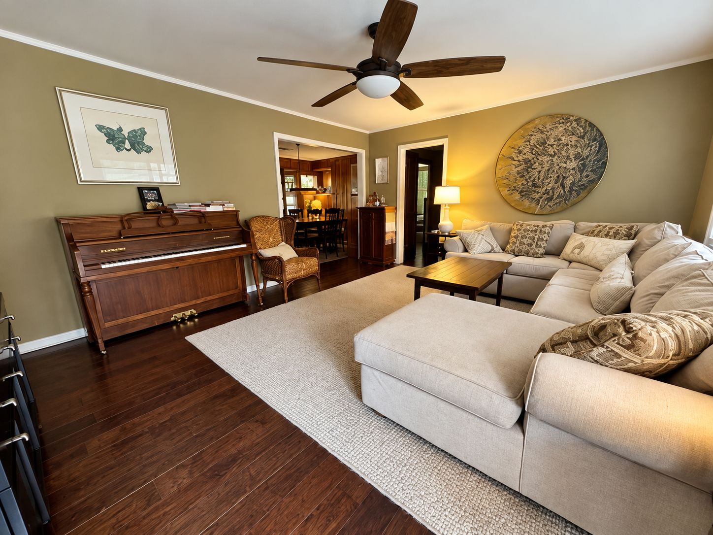 A living room with a piano, a sofa, a coffee table, a ceiling fan, and a dining area in the background.