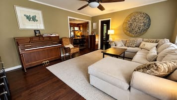 A living room with a piano, a sofa, a coffee table, a ceiling fan, and a dining area in the background.