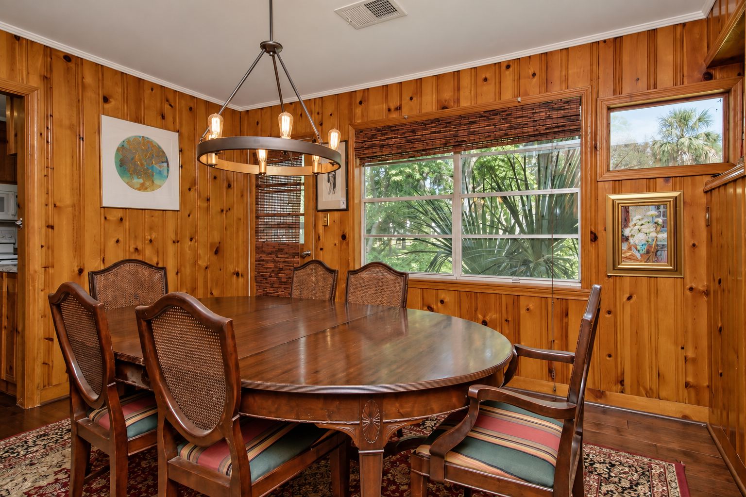 A dining room with a wooden paneled wall, a round table, six chairs, a chandelier, and large windows with a view of greenery.