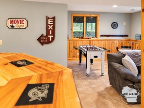 A room with a foosball table, a sofa, and a wooden table with a logo. There's a window with a view of trees and a clock on the wall.