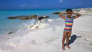 A person standing on a sandy beach with clear turquoise water and rocky formations in the background.