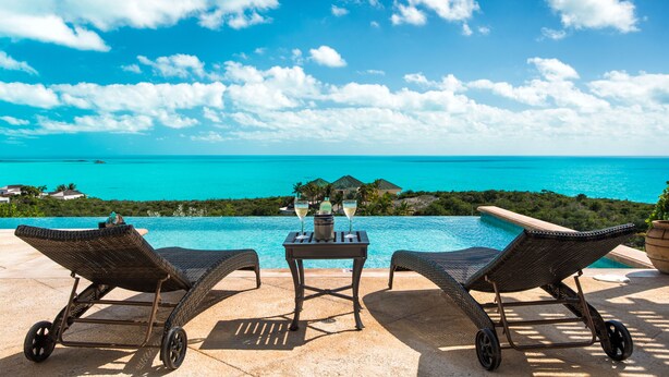 A poolside area with two wicker lounge chairs, a table with two glasses of champagne, and a view of the ocean and distant buildings.