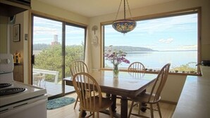 Un espace repas comprenant une grande table et des chaises, un vase de fleurs et une vue sur une étendue d’eau, visible à travers de grandes fenêtres.