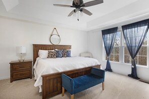 Quarto com cama de madeira, mesa de cabeceira, um banquinho, ventoinha no teto e janela com cortinas.