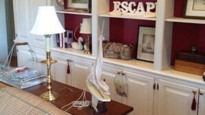 A room with a wooden table, a lamp, a white swan sculpture, and a bookshelf with various decorative items and a sign that reads "ESCAP". -  Cottage on a Private Island in the York River (Gloucester Point)