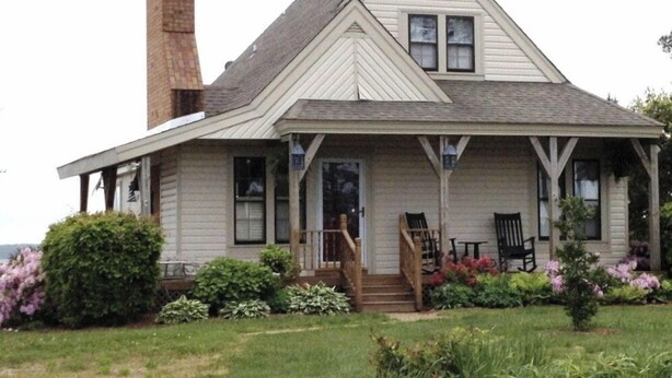 A two-story house with a prominent brick chimney, a front porch with steps, and a small garden with various plants. -  Cottage on a Private Island in the York River (Gloucester Point)